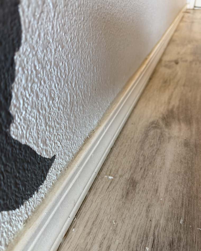 Hallway baseboard before cleaning, with dust built up along the trim and debris on the floor